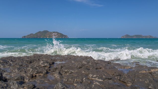 Waves Crashing on Rocky Shoreline with Islands in Background Under Clear Blue Sky