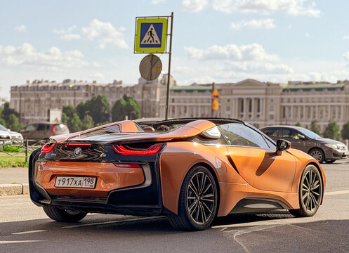 An orange luxury BMW i8 Roadster sports car driving on a city street road against historical buildings and a pedestrian crosswalk sign.