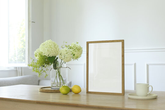 Blank Wooden Picture Frame Poster Mockup. Glass Vase with Hydrangea Flowers, Lemons Fruit and Coffee Cup on Table. Open Window in Bright Scandinavian Interior. Home Decor Background. Natural Light