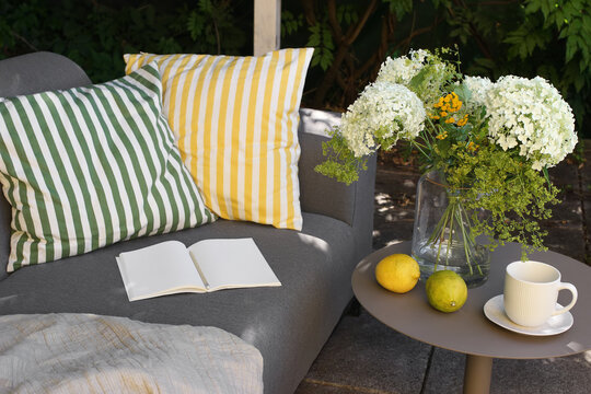 Cozy Outdoor Patio Lounge with Yellow Green Striped Cushions, open notebook in sunlight. Vase with Hydrangea, Tansy, Alchemilla Flowers. Lemon fruit, Coffee Tea Cup on side table. Summer relaxation.