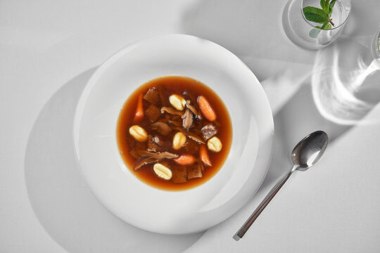 Gourmet mushroom broth soup with gnocchi and baby carrots on a white plate. Minimalist top view fine dining table setting with hard shadows.