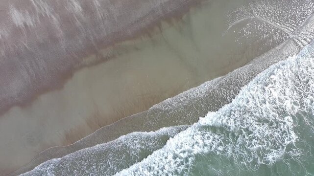 30 second aerial footage from a drone looking down as a group of waves gently wash up on a sandy beach. 