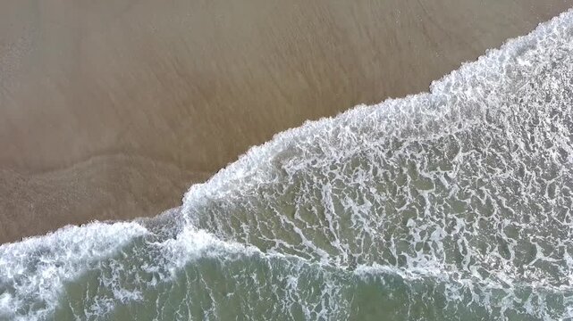 30 second aerial footage from a drone pulling up as it looks down on waves gently crashing on a sandy beach. 