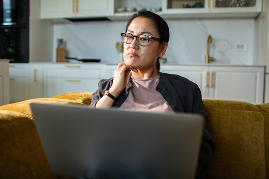 Thoughtful woman with glasses working on laptop at home kitchen