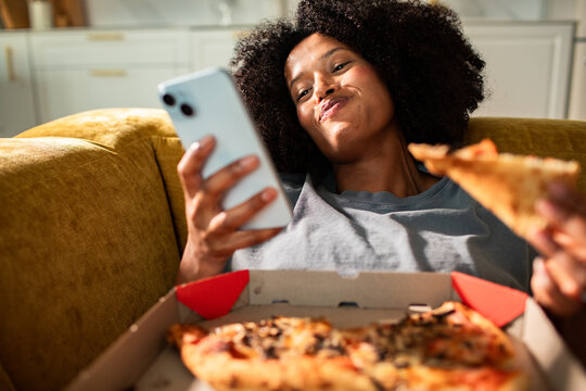 Young woman eating pizza and texting at home on sofa