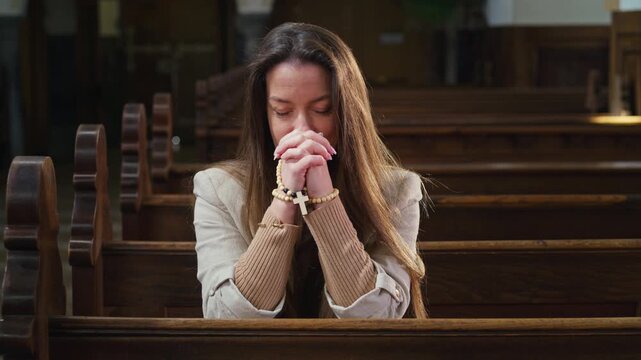Devout woman sitting in prayer in a church pew, with eyes closed and clasped hands, holding a rosary beads with a cross.