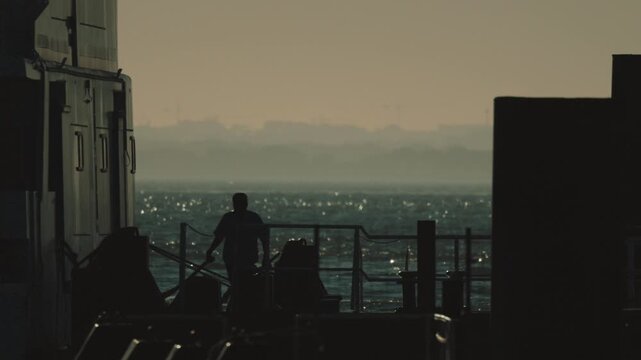 Male worker on a ship deck adjusts equipment while silhouetted against the ocean, with distant land visible on the horizon during sunset