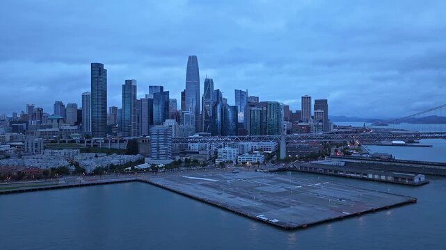 Drone video glides over San Francisco&rsquo;s modern skyline and waterfront at dusk, capturing iconic Salesforce Tower, moody blue lighting, dramatic clouds, and glass city architecture.