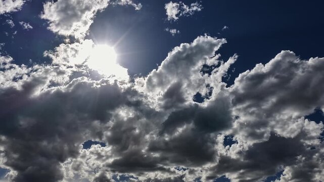 4K UHD steady timelapse of a deep, vivid blue sky pierced by the bright sun. Fast-moving, highly contrasted silver and chrome-grey clouds shift dynamically across the wide frame.