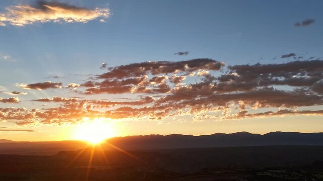 4K UHD drone timelapse of a golden New Mexico sunrise. The bright sun crests over a dark mountain silhouette, casting sharp downward sunbeams through a layer of scattered, textured morning clouds.