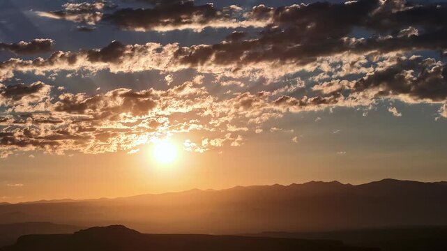 4K UHD drone timelapse of a brilliant golden sunrise. The glowing sun ascends from behind a dark Southwestern mountain ridge, illuminating a textured layer of morning clouds in a vibrant orange sky.
