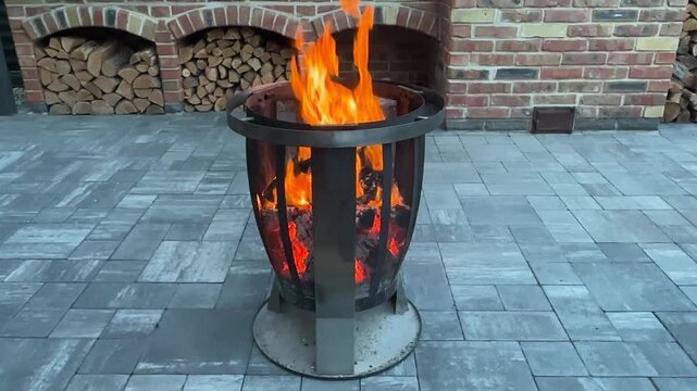 Enjoying fire in a metal fire pit on a patio with bricks and stacked wood in the background during evening hours