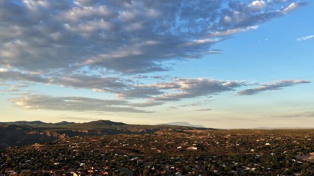 4K UHD drone timelapse capturing soft dawn light over a suburban canyon town. Fast moving, textured clouds pass across a blue sky over the sprawling neighborhood, plateau, and distant mountains.