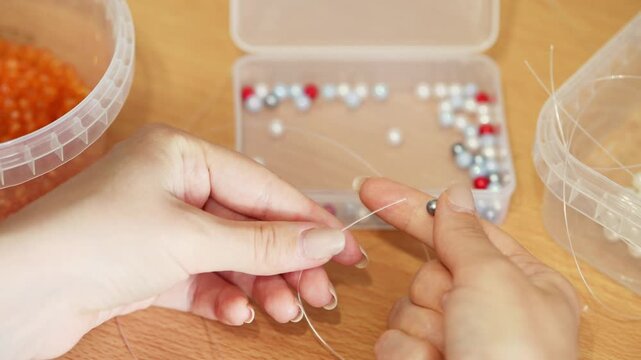 Hands threading beads with translucent thread on wooden table in natural light