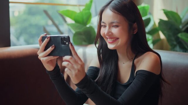 Happy Asian woman smiling while holding smartphone horizontally to watch online streaming video or playing mobile games in cafe, young female creator using phone for entertainment, digital lifestyle