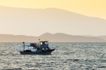 Obraz premium Traditional small fishing boat sailing on calm sea water during a hazy sunset with silhouettes of distant mountains and islands in the background.