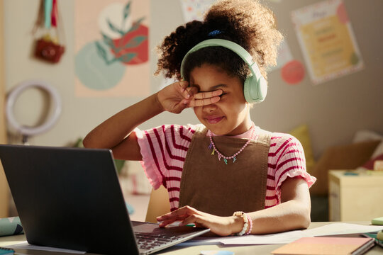 African American child wearing headphones rubbing eyes while using laptop for online learning, showing screen fatigue and study overload. Useful for edtech or parenting campaigns