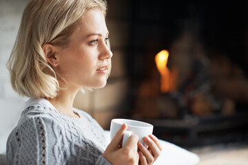 Obraz premium Fireplace, home and woman with coffee, thinking and contemplating decision with hot drink in lounge. Remember, memory and person with warm beverage on weekend, space and reflection with tea in house