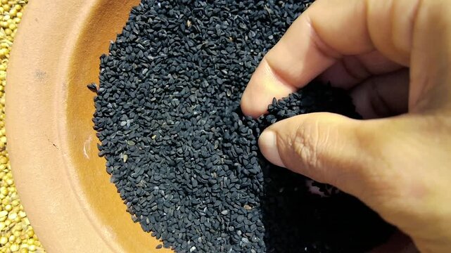 A close up of a person's hand picking up a handful of black cumin seeds from a clay bowl