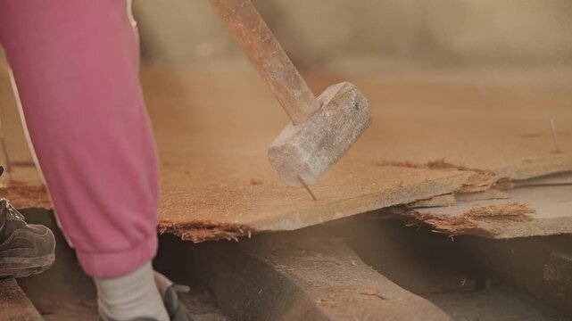Worker bends stubborn nail while restoring worn wooden floor in dusty apartment