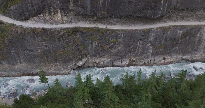 Annapurna CIrcuit, Manang region, Nepal, tracking drone shot of a lone hiker trekking on a high cliffside trail above the rushing Marsyangdi River and pine forests.