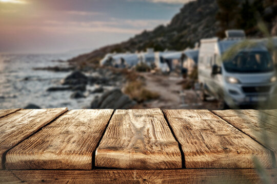 Copy space on empty wooden table with the background of camping scene with camper van by rocky seaside &ndash; summer travel and outdoor adventure concept.