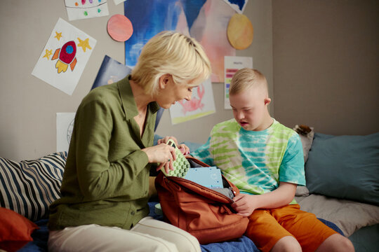 Caucasian mother helping boy with Down Syndrome packing school backpack, preparing learning supplies at home. Education support concept, useful for parenting, inclusion, caregiving campaigns