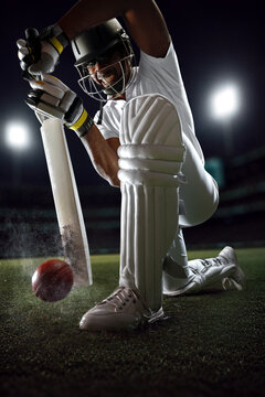 Low-angle cricket batter blocking red ball beside protective leg pads under stadium lights. Concept of batting defense, player protection, cricket competition, sports branding and promotion.