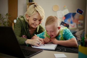 Caucasian mother helping child with Down Syndrome writing in notebook at home, supporting learning...
