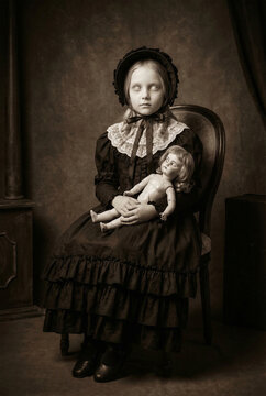 Eerie vintage sepia studio portrait of a creepy possessed little girl with blank white eyes in a dark Victorian dress and bonnet sitting on an antique chair holding a spooky old doll