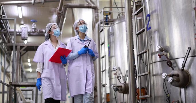 Female engineers inspecting industrial storage tank and piping system in food factory, Quality control specialists auditing production standards near large stainless steel vats in industrial plant. 