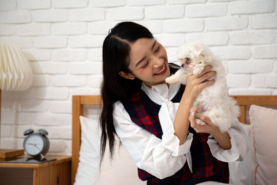 Smiling young woman holding a white puppy dog in a cozy bedroom, sharing affection, happiness, love, care, companionship, tenderness, playfulness, and joyful bonding at home