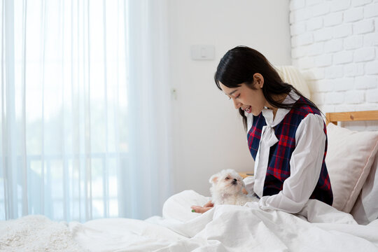 Smiling young woman sitting on a bed with a white puppy dog, sharing affection, care, companionship, comfort, happiness, and playful bonding in a bright cozy bedroom at home