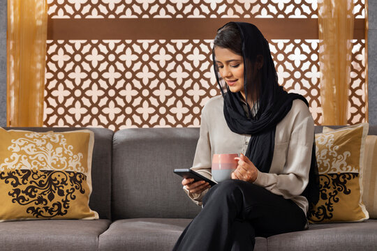 Muslim woman using mobile phone while drinking coffee at home