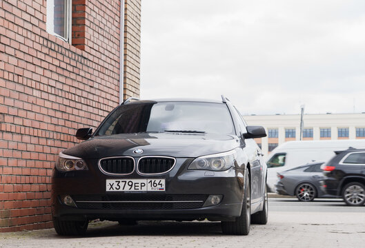 Front view of a tuned dark gray BMW 5 Series E61 LCI facelift station wagon car with an M Sport front bumper and black kidney grilles, parked on a street sidewalk near a brick wall.