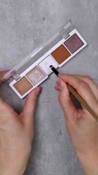 A woman's hands test an eyeshadow palette with a brush on the skin of her hands on a table.