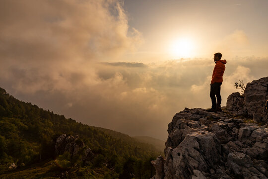 Hiker in orange jacket gazing at sunset over clouds from mountain cliff for outdoor adventure, Monte Baldo Natural Park, Brentonico, Trentino, Veneto, Italy.