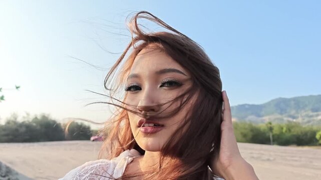 Close-up of a young Indonesian southeast asian woman in a white lace dress posing on a windy sand dune. Her hair blows across her face as she touches it. Sandy dunes and hills under a clear blue sky