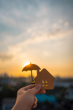 Hand holding a wooden house cut-out with an umbrella silhouette against sunset. Concept of home insurance, financial protection, property safety, and mortgage security for family.