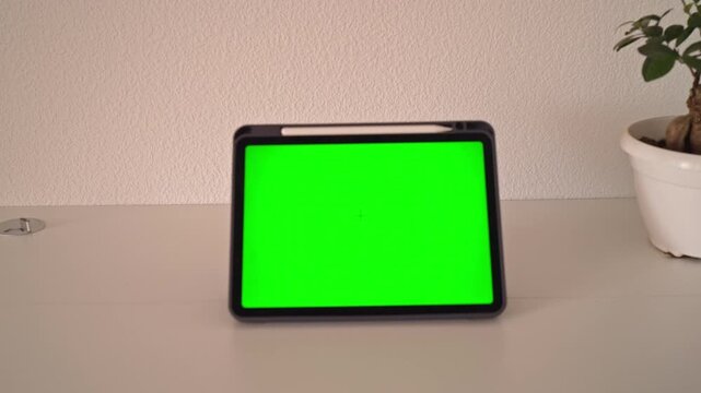 A tablet is placed on a table with a green screen visible. There is a small plant in a pot next to the tablet. The space is simple and well lit.