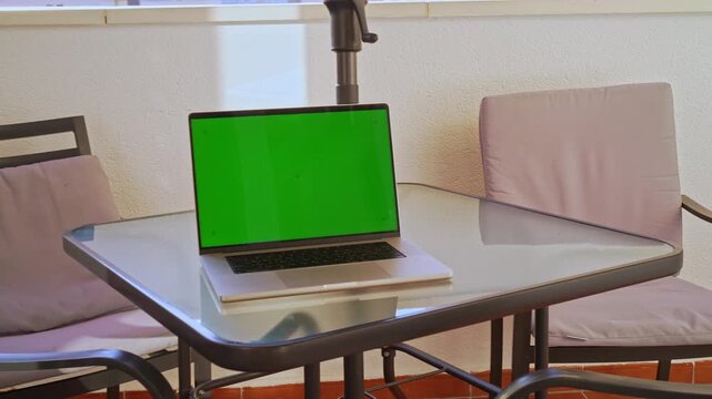 A laptop with a green screen sits on a table at an outdoor balcony. The sunlight shines on the furniture. The setting is simple with a few chairs and a clear view.