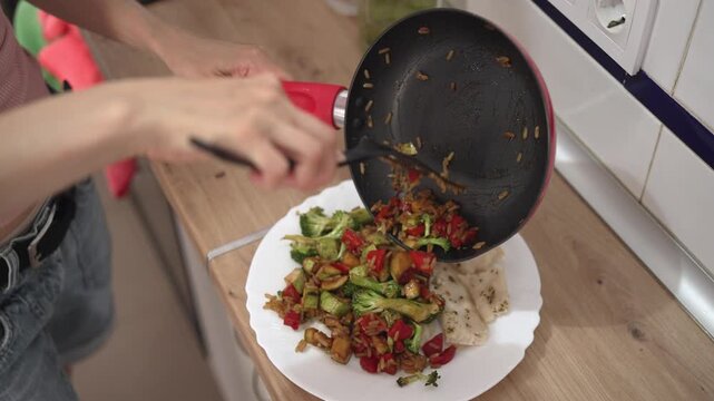 A person cooks vegetables in a pan and then serves the stir-fry onto a plate with rice. The kitchen has bright light and clean surfaces. Food preparation shows simple cooking methods.