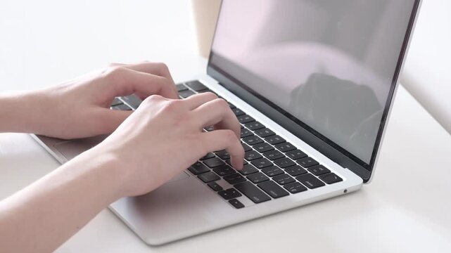 Hands typing on open laptop with blank bright screen in minimalist workspace, showing steady keyboard use on white desk with soft neutral light, clean modern composition for office tasks and online