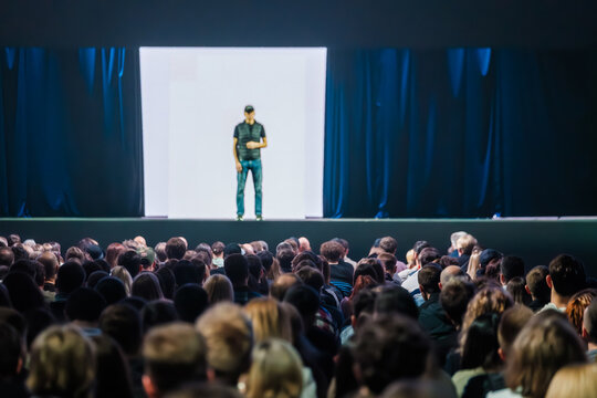 Large audience focused on lone speaker delivering presentation on illuminated stage, dramatic lighting and attentive crowd in conference auditorium.