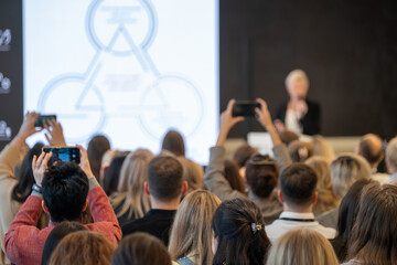 Obraz premium Crowd at business conference recording keynote presentation with smartphones, engaged audience, networking, learning.