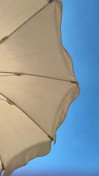 Slow pan shot of beach umbrella canopy gently swaying across clear blue sky with scalloped edge visible handheld tilt revealing fabric ribs over seaside horizon at midday