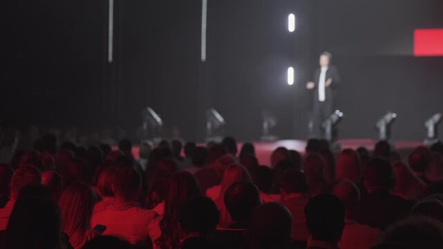 Audience watching keynote speaker on stage at corporate conference. Dramatic red lighting creates moody, focused atmosphere.