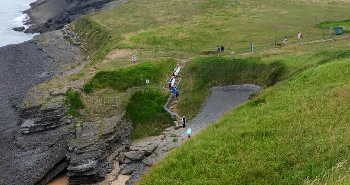 Coastal Trail View: A scenic view of a coastal path with grassy slopes leading down to rocky shores and the ocean. Several people are seen walking and enjoying the outdoors.