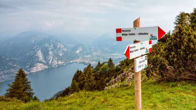 Hiking signpost indicates mountain paths with a scenic view over Lake Garda and Riva town in a summer alpine adventure destination, Monte Baldo Natural Park, Brentonico, Trentino, Veneto, Italy.