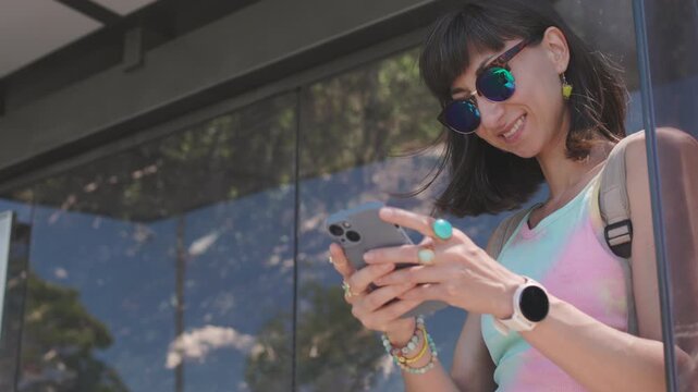 A young woman stands at a modern bus stop, holding a smartphone. She smiles and glances upward while touching the screen and scrolling. her hair is gently blowing in the breeze. A vibrant cityscape.